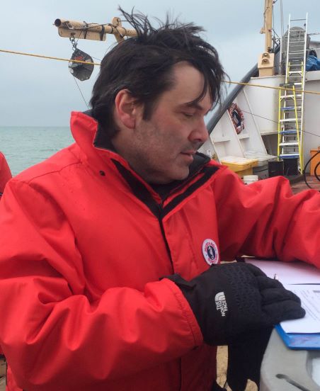 Dr. McKay on a ship writing notes wearing an orange jacket