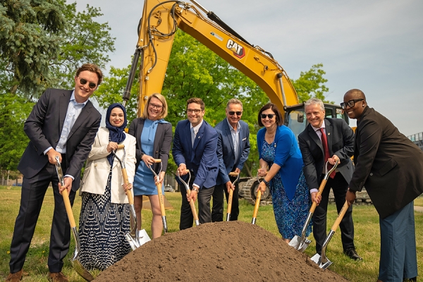 Line of people holding shovels before pile of soil.