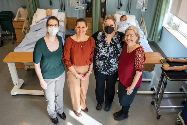 Four women stand at foot of hospital pateint&#039;s bed: Dana Ménard, Laurie Freeman, Kendall Soucie, and Jody Ralph