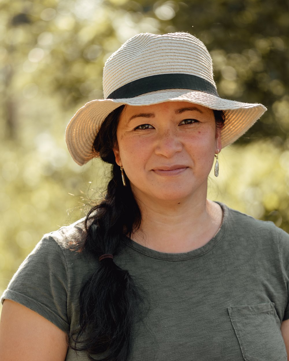 Photo of a woman with long dark hair, standing in front of trees. She wears a sraw hat