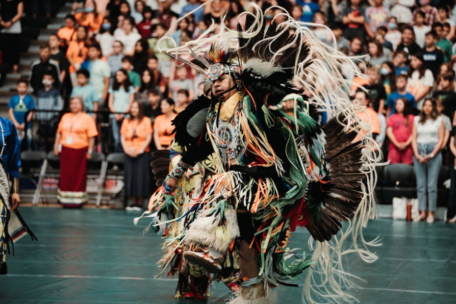 An Indigenous male dancer at a pow wow dances in a brightly coloured regalia, adorned with feathers, beads, and intricate designs. He is mid-step, one foot lifted off the ground, and his arms are extended out to his sides. In the background, spectators ca