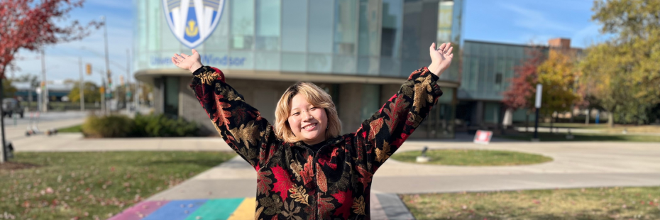 Fei standing on the Rainbow Crosswalk in front of the Welcome Centre smiling at the camera with their arms up in the air